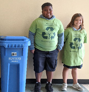 Two students next to recycle bin wearing recycle t-shirts